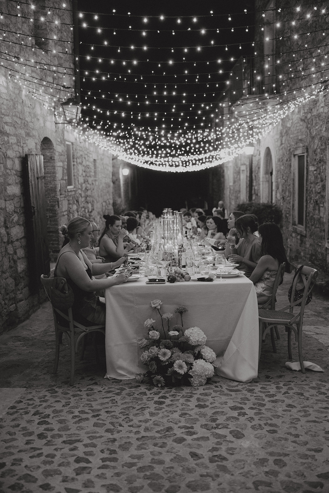 Dinner along the stone street of Borgo Laticastelli beneath a canopy of fairy lights at night, photographed in black and white